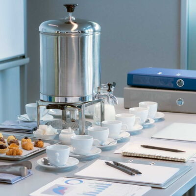 Konferenztisch mit mehreren Kaffeetassen, Gebäck, einem großen silbernen Getränkespender, Schreibutensilien und zwei Aktenordnern