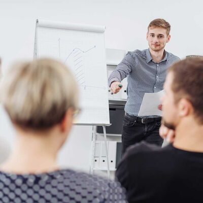 Ein Mann steht vor einer Flipchart mit Diagramm und spricht zu mehreren sitzenden Personen.