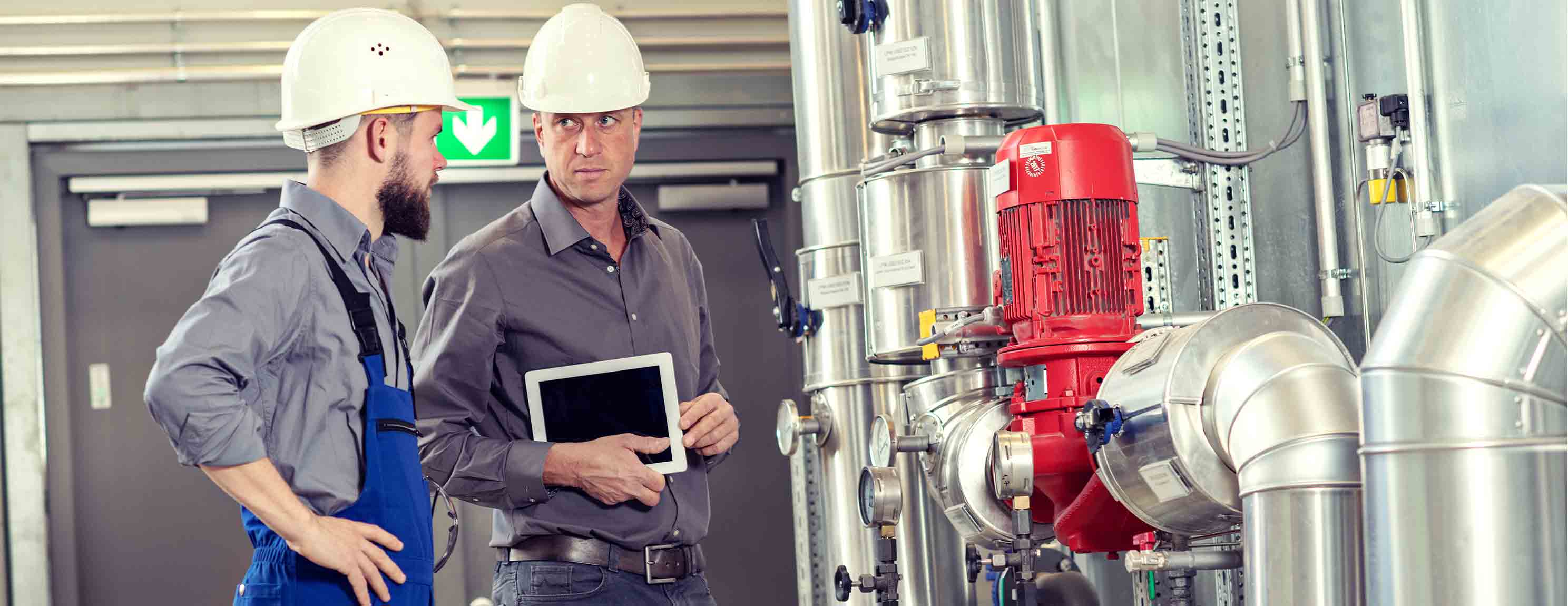 Zwei Männer mit weißen Helmen diskutieren vor industriellen Maschinen. Einer hält ein Tablet, der andere trägt blaue Arbeitskleidung. Hintergrund zeigt metallene Rohrleitungen und ein rotes Gerät.