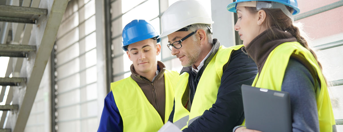 Drei Personen stehen mit gelben Schutzwesten und Schutzhelmen stehen nebeneinander in einer Halle und schauen sich Pläne an.