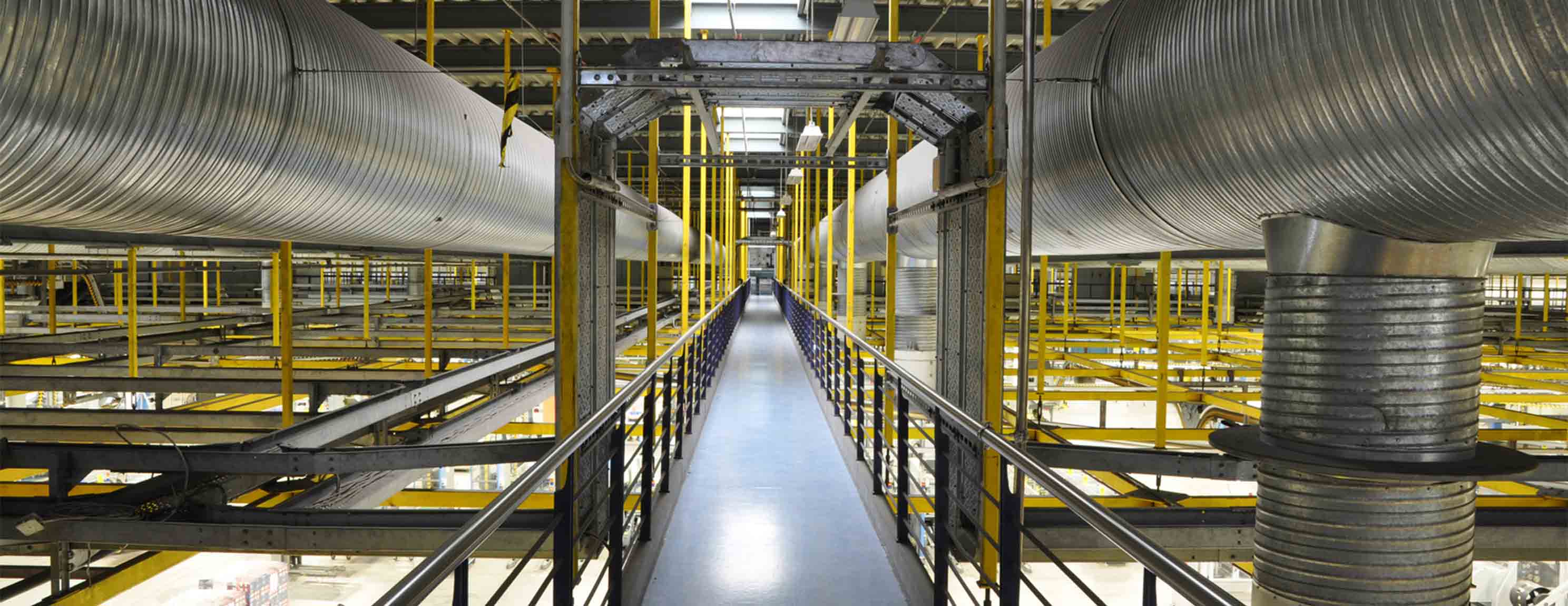 Ein langer Metallgang erstreckt sich durch eine Industriehalle. Große Lüftungsrohre verlaufen parallel darüber. Gelbe und schwarze Trägerstrukturen sichern die Umgebung.