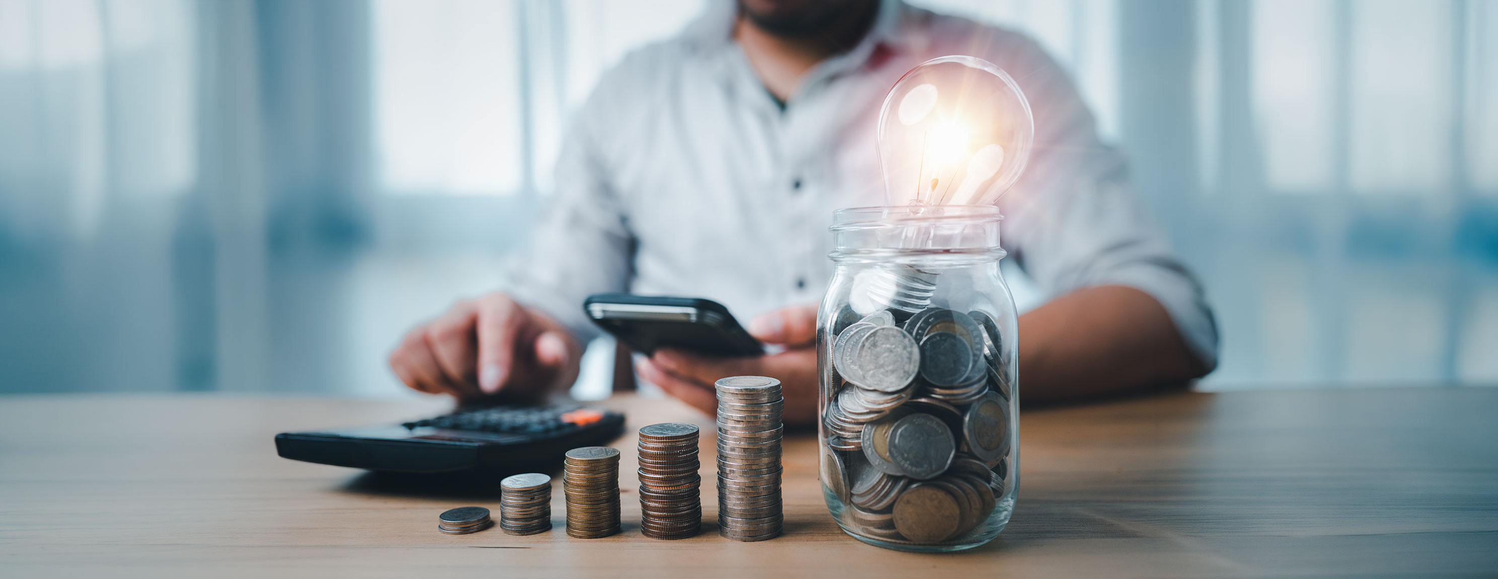 Hausmeister mit Smartphone, daneben Münzglas und leuchtende Glühbirne, Thema Energiesparen.