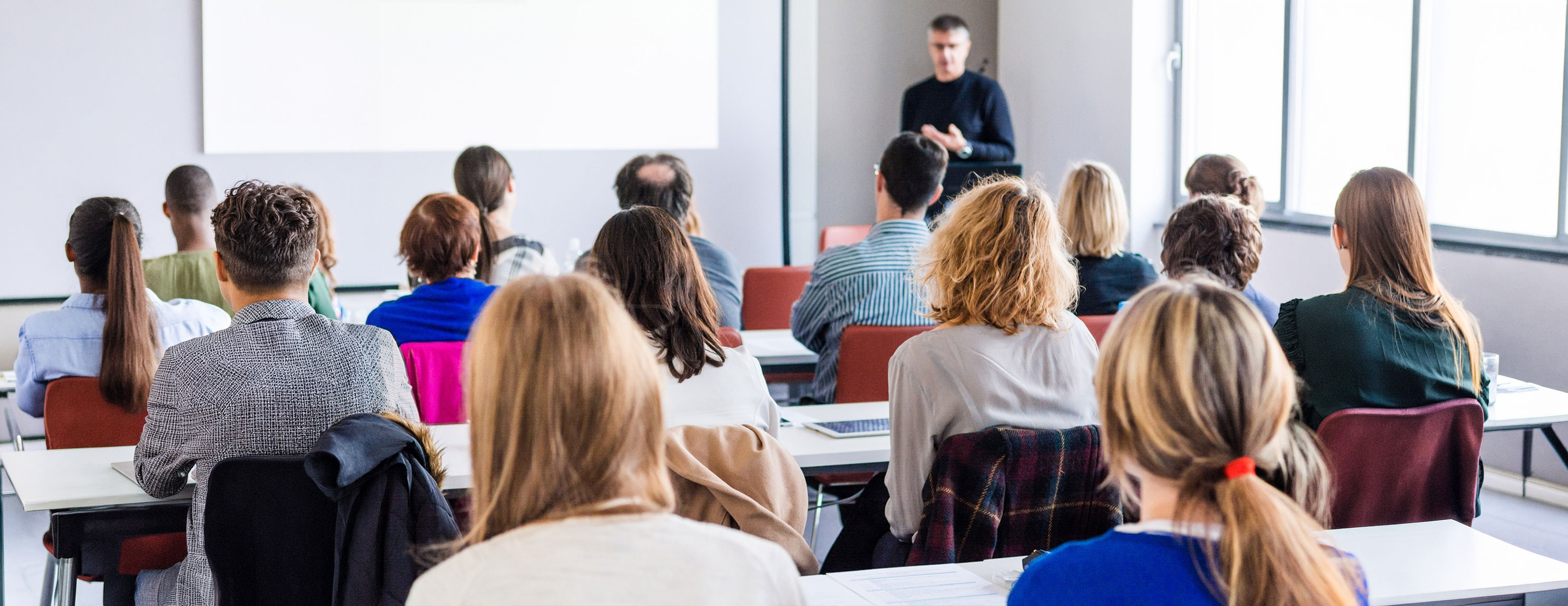 Gruppe von Menschen sitzt in einem hellen Schulungsraum und hört einem stehenden Vortragenden zu